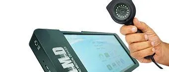 A hand holds a small round camera with LED lights next to a portable video monitor labeled “MD” on its casing.