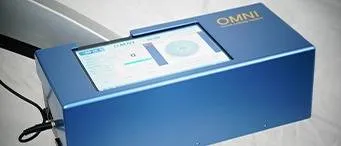 A blue rectangular device labeled OMNI with a touchscreen display showing data and charts, connected by a cable on one side, placed on a light-colored surface.