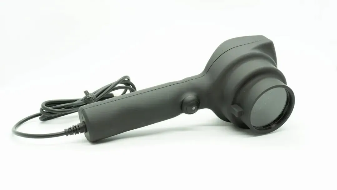 A black handheld device with a round lens at the front, a power button on the handle, and an attached power cord, shown against a plain white background.