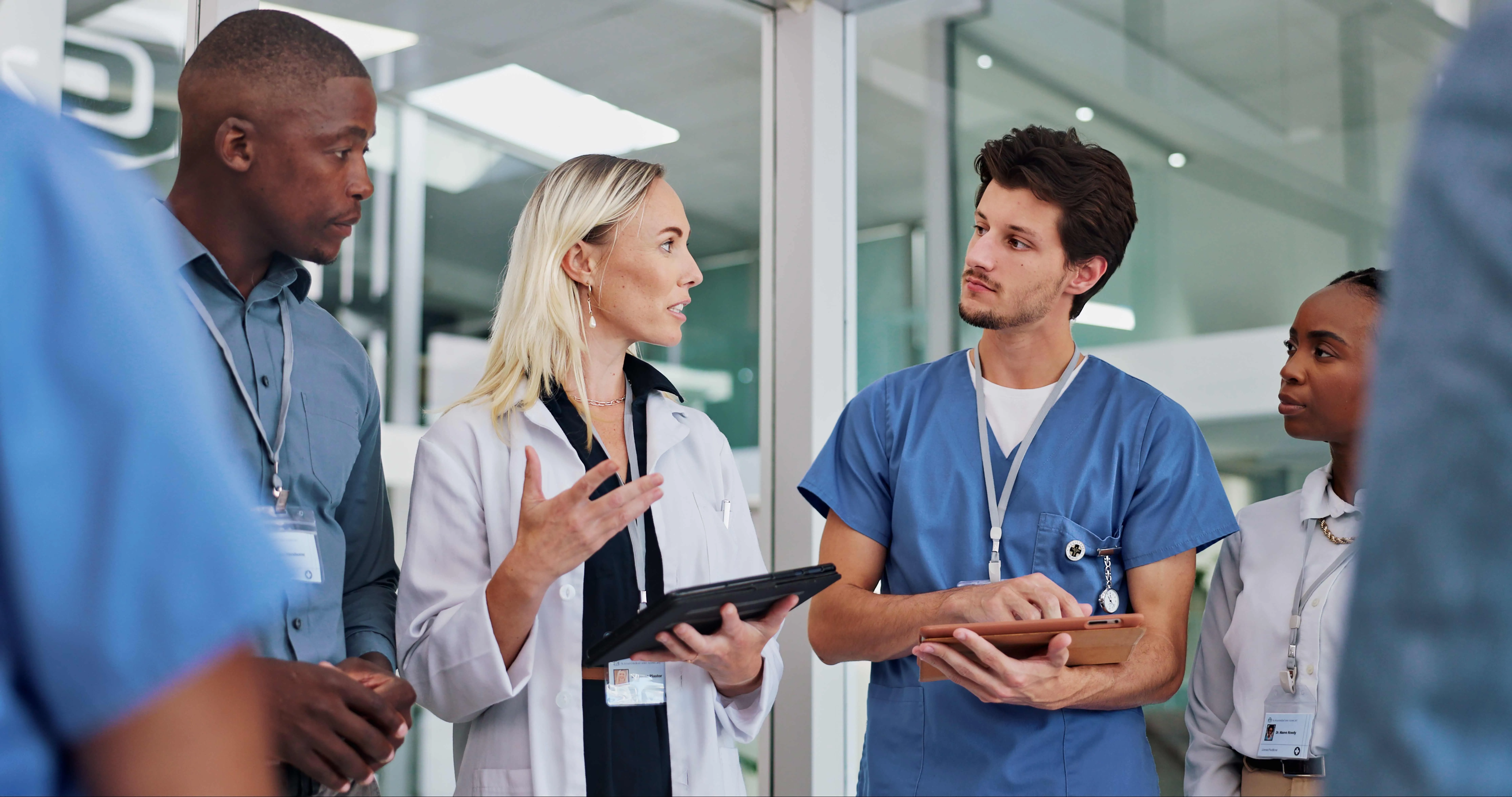 Four healthcare professionals, including doctors and nurses, stand together in discussion, holding tablets and wearing medical attire, inside a modern hospital or clinical setting.