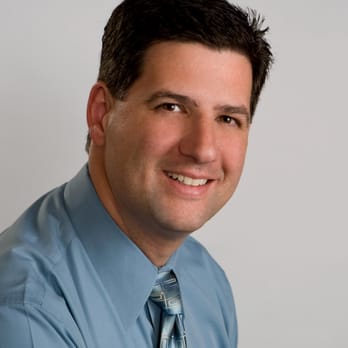 A man with short dark hair wearing a light blue dress shirt and patterned tie is smiling while posing for a professional headshot against a plain light background.