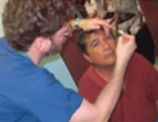A healthcare professional in blue scrubs examines an older man’s eye using a medical instrument, while the man in a red shirt looks upward. Other people are visible in the blurred background.
