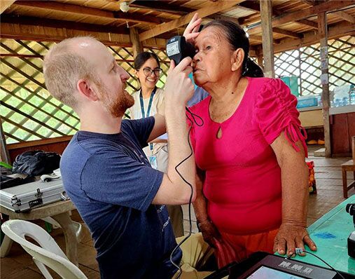A man examines an older woman’s eye using a handheld device in a wooden building; the woman stands while another woman in the background smiles, observing the scene.