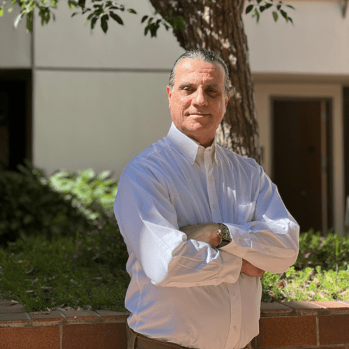 A man in a white dress shirt stands outdoors with his arms crossed, in front of a tree and some greenery, with sunlight shining on him and a building in the background.