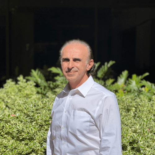 A man with light skin and thinning hair stands outdoors in front of green leafy bushes, wearing a white button-up shirt and looking at the camera, with sunlight illuminating his face and shirt.