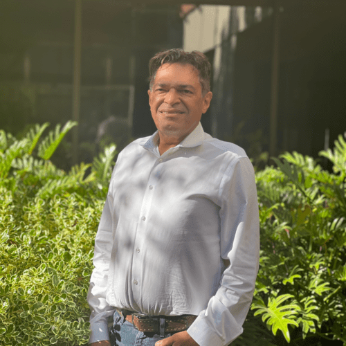 A man wearing a white button-up shirt stands outdoors in front of lush green plants, smiling at the camera. Sunlight casts soft shadows on his face and the foliage behind him.
