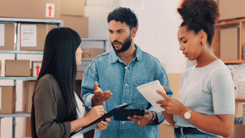 Three people stand in a warehouse talking. One man holds a tablet and gestures, while two women listen; one holds a pen and notepad. Boxes are stacked on shelves in the background.