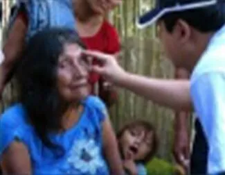 A person in a cap touches the forehead of an older woman in a blue shirt, while a child and two other adults stand nearby in an outdoor setting.