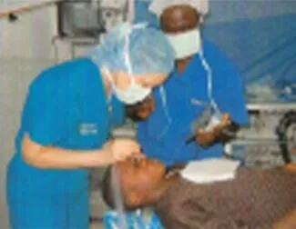 Two medical professionals wearing scrubs, masks, and hair covers perform an eye procedure on a patient lying on a table in a clinical setting.