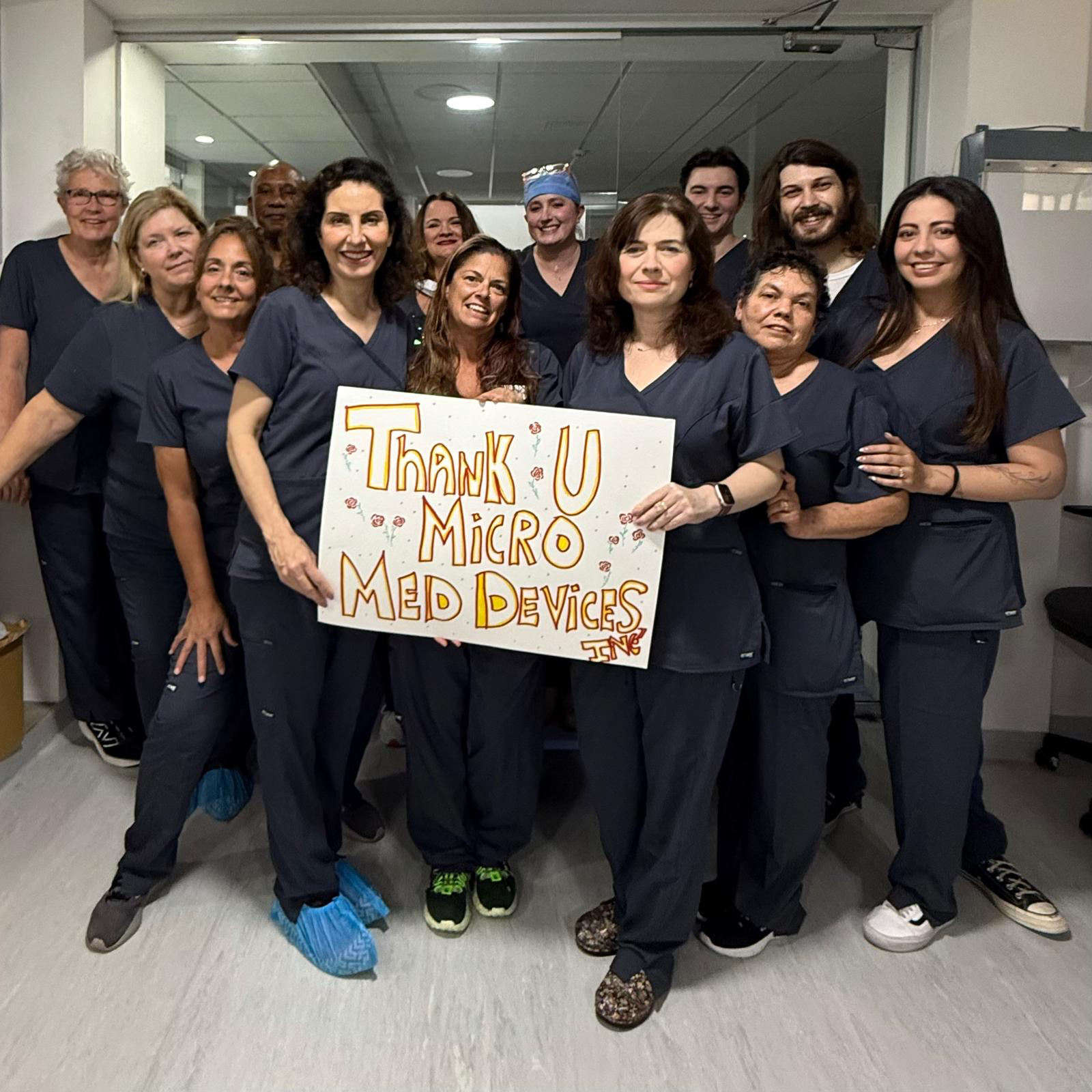 A group of smiling medical staff in scrubs stand together in a hallway, holding a colorful sign that reads Thank U Micro Med Devices.