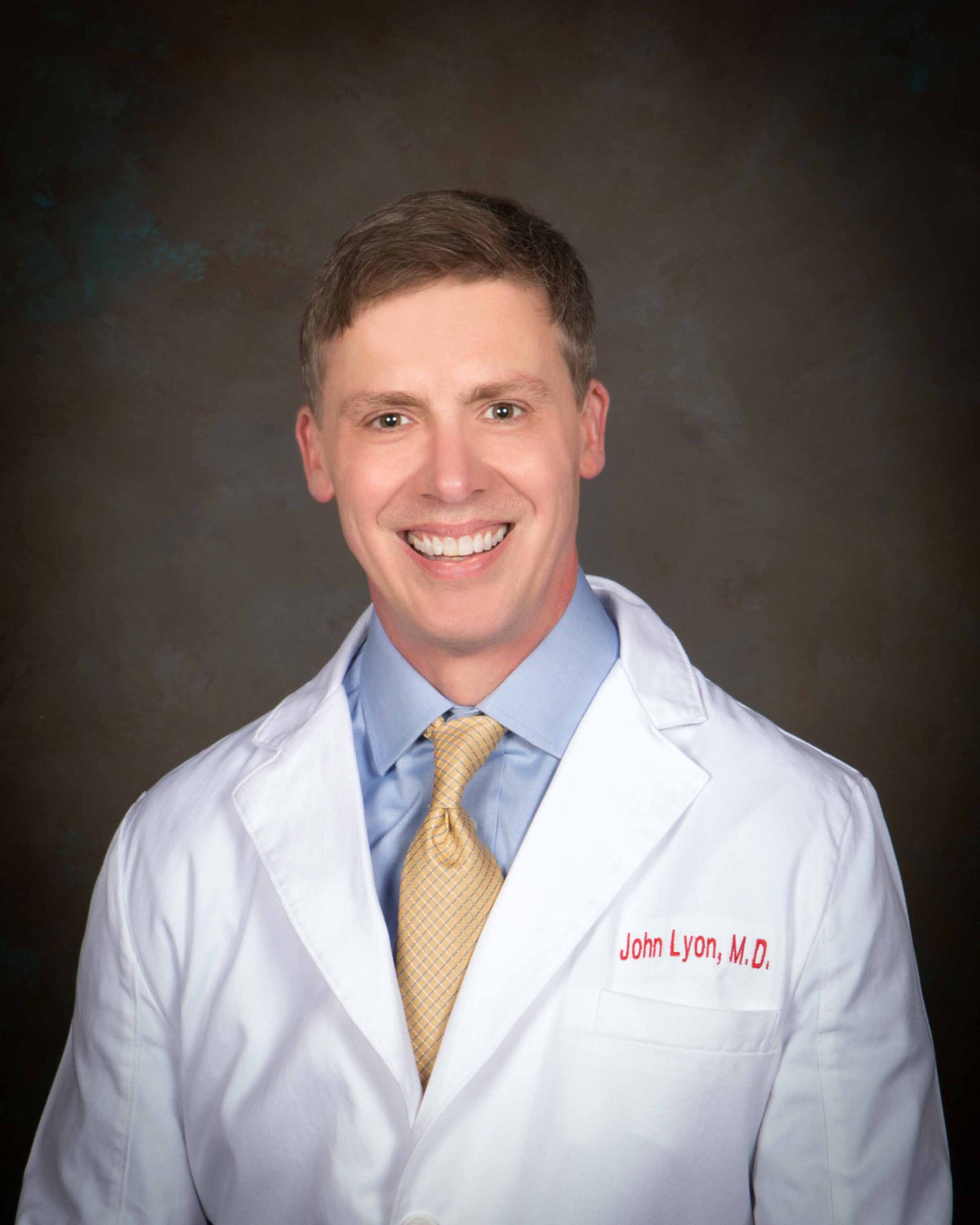 A smiling man wearing a white lab coat embroidered with John Lyon, M.D., a blue shirt, and a yellow tie, poses against a dark, neutral background.