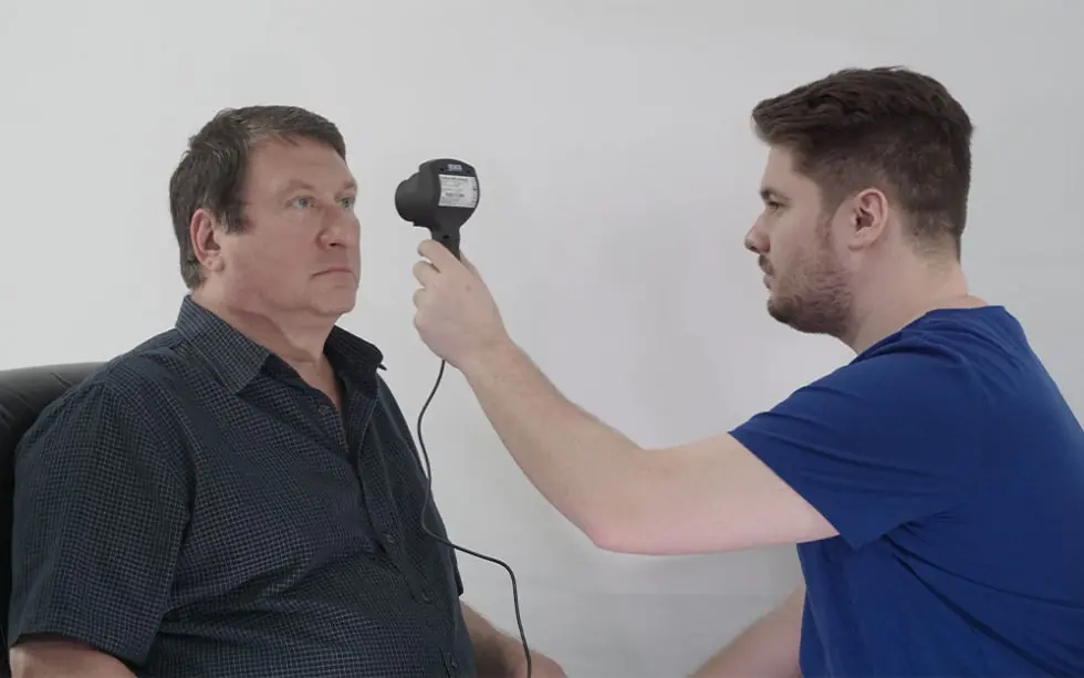 A man sits while another man in blue holds a handheld medical device close to his left eye, likely performing an eye examination against a plain white background.